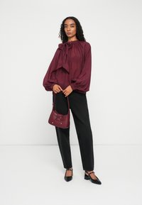 Bluză burgundă cu guler tip fundă, mâneci balloon, asortată cu pantaloni negri tapered și o geantă crossbody maronie cu detalii în fundă. Tocuri negre.