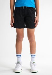 Pantalones cortos negros con cintura de cordón, que quedan por encima de la rodilla. Combinados con calcetines blancos con rayas azules y zapatillas blancas.