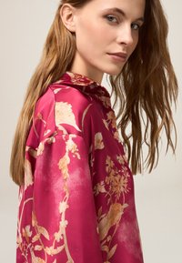 Blusa in seta floreale di un fucsia vivace, con colletto, vestibilità morbida e intricato motivo floreale in oro e crema. Texture liscia, design fluido.