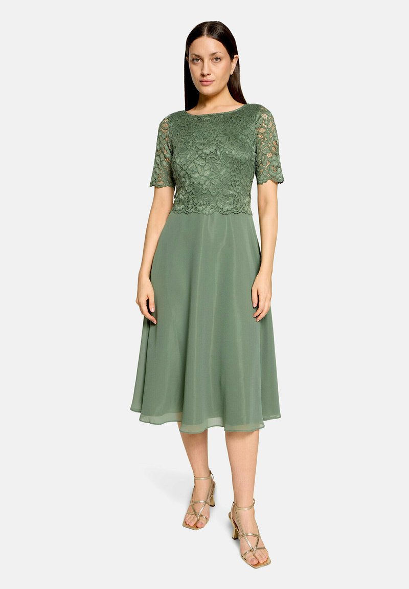 Femme portant une robe vert olive longueur genou avec manches courtes en dentelle et corsage, associée à des sandales dorées à talons hauts à brides.