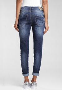 Žena v tmavě modrých skinny džínech s ohrnutými nohavicemi a stříbrných teniskách Adidas, zobrazená zezadu na jednolité šedé pozadí.