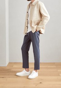 Beige arbetsjacka med svarta knappar, vit T-shirt, marinblå slim-fit byxor och vita sneakers, mot en ljus bakgrund.