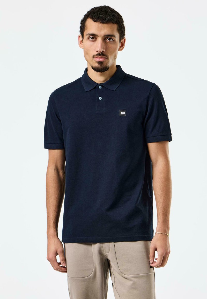 Mörkblå polo t-shirt i bomull med knappad krage, korta ärmar och en liten logotyp patch på vänster bröst.