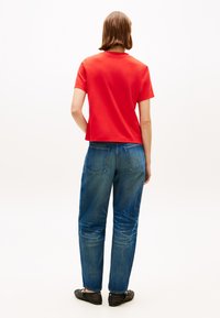 Κόκκινο βαμβακερό μπλουζάκι με κοντά μανίκια, συνδυασμένο με μπλε τζιν παντελόνι κανονικής γραμμής. Το σύνολο ολοκληρώνεται με μαύρα παντόφλες με τρουκς.