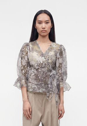 Mladá žena nosí priesvitnú kvetinovú zavinovaciu blúzku s nadýchanými rukávmi a béžové nohavice s vysokým pásom na bielom jednoduchom pozadí.