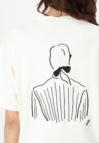 Cream cotton t-shirt featuring a black line drawing of a figure with a bow, displaying vertical stripes on the back design.