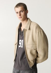 Beige velúr bomber dzseki, cipzárral, két elülső zsebbel és bordás mandzsettával. Sötétszürke póló fölött viseli, amelyen fehér "91." felirat található.