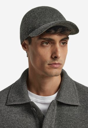 Grå ullkeps med rund krona och en styv, något böjd skärm. Modellen bär en grå jacka med krage över en vit skjorta.