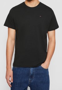 Camiseta de algodón negra con mangas cortas y cuello redondo, que presenta un pequeño logo tricolor en el pecho. Lleva puesta unos pantalones vaqueros de mezclilla azules.