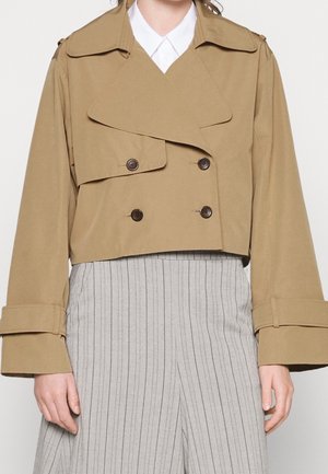 Gabardina corta beige de doble botonadura con solapas anchas y correas en los puños, llevada sobre una camisa blanca y pantalones grises con rayas finas.