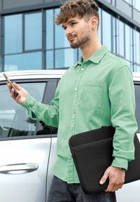 Mann in grünem Hemd, der ein Smartphone und eine schwarze Laptoptasche hält, steht neben einem weißen Auto in städtischer Umgebung.
