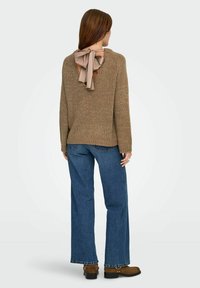 Suéter de punto beige con un detalle de lazo en la parte posterior, combinado con jeans de pierna ancha azules y botines marrones. Textura suave y ajuste relajado.