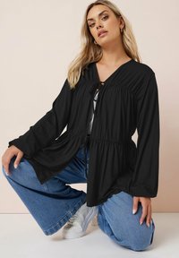 Blusa nera a maniche lunghe con scollatura a V e chiusura con fiocco, caratterizzata da tessuto arricciato in vita, abbinata a jeans larghi azzurri e sneakers bianche.