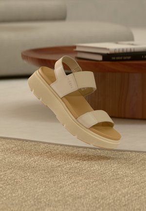 Bézs nyitott orrú szandál vastag talppal és állítható szövetpántokkal, amely egy modern nappaliban, egy texturált szövött szőnyeg felett lebeg.