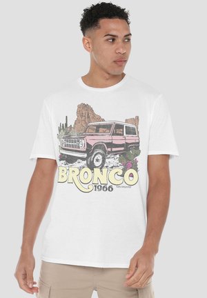 Young man wearing a white T-shirt with a vintage pink Bronco 1966 vehicle, desert cacti, and rock formations graphic.