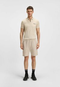 Polo beige con zip e un motivo sottile, abbinato a pantaloni corti beige. Calzini neri con logo e sneakers nere completano l'outfit.