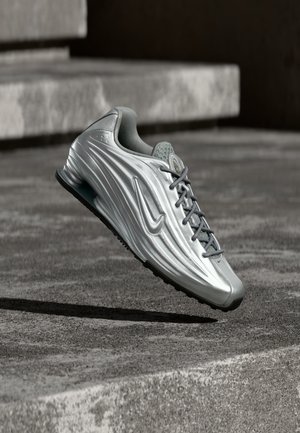 Srebrny sneaker Nike z teksturowanym wzorem po bokach i szarymi sznurowadłami unoszący się nad betonowymi schodami na stonowanym outdoorowym tle.