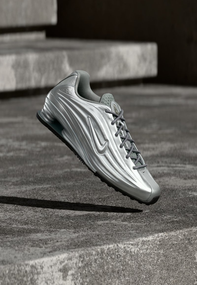 Ασημί αθλητικό παπούτσι Nike με υφή στο πλευρικό σχέδιο και γκρι κορδόνια, αιωρείται πάνω από σκάλες από σκυρόδεμα σε ήπιο υπαίθριο περιβάλλον.