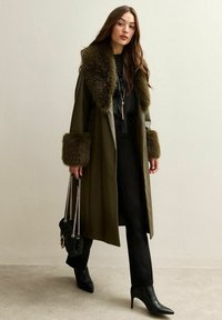 Oliwkowy płaszcz z futrzanym kołnierzem i mankietami, zestawiony z czarnymi ubraniami i spiczastymi botkami za kostkę. Czarna torebka z łańcuszkowymi paskami.