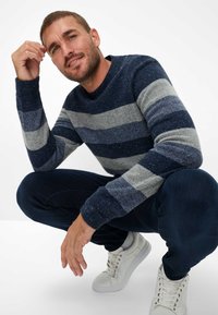 Maglione lavorato a maglia con strisce orizzontali in blu navy, grigio e azzurro chiaro, con polsini a coste e scollatura rotonda, indossato con jeans scuri e sneakers bianche.