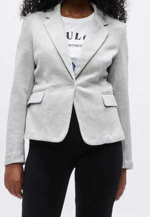 Kvinde iført en lysegrå enkelknappet blazer over en hvid T-shirt med grafik og sorte bukser.