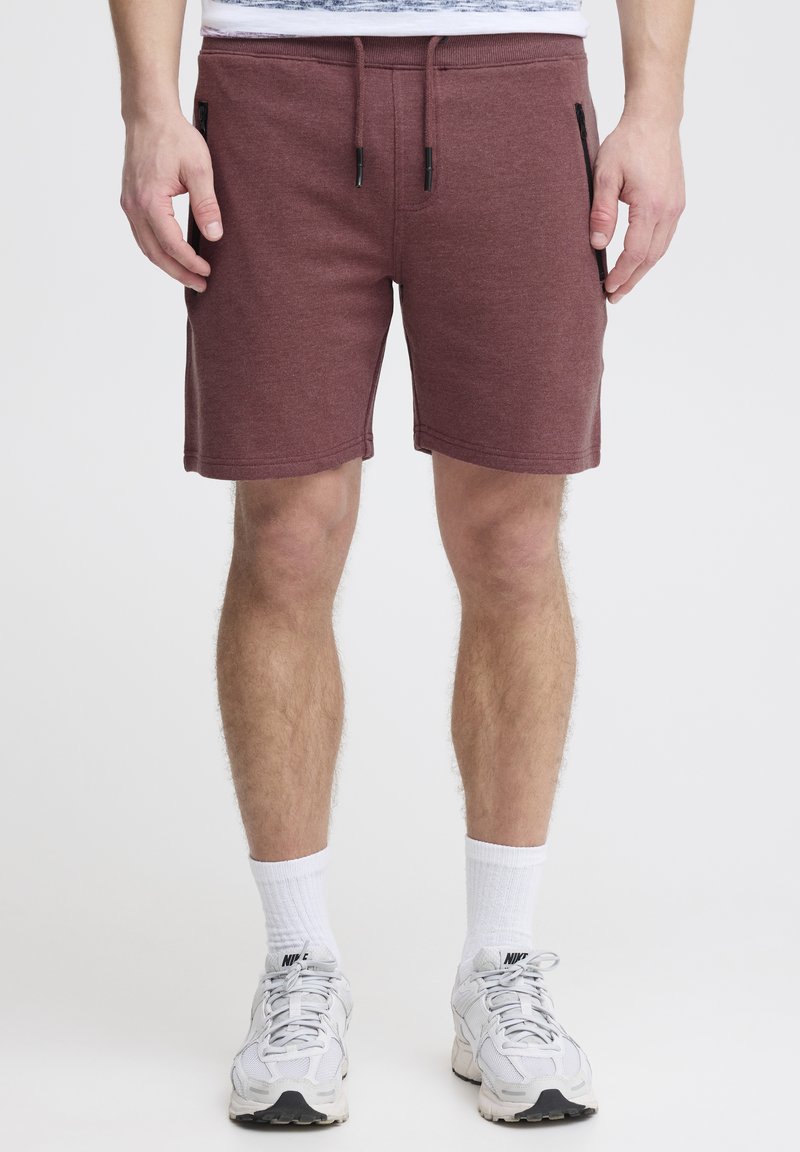 Shorts bordeaux avec une taille à cordon de serrage, et deux poches latérales zippées noires. Fabriqués dans un tissu texturé, se terminant au-dessus du genou.