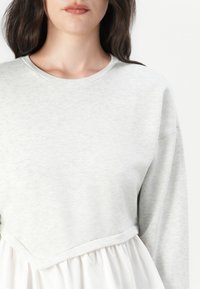 Top de manga larga gris con cuello redondo, que presenta un dobladillo acampanado blanco en contraste. Textura suave, corte relajado y diseño sin costuras.