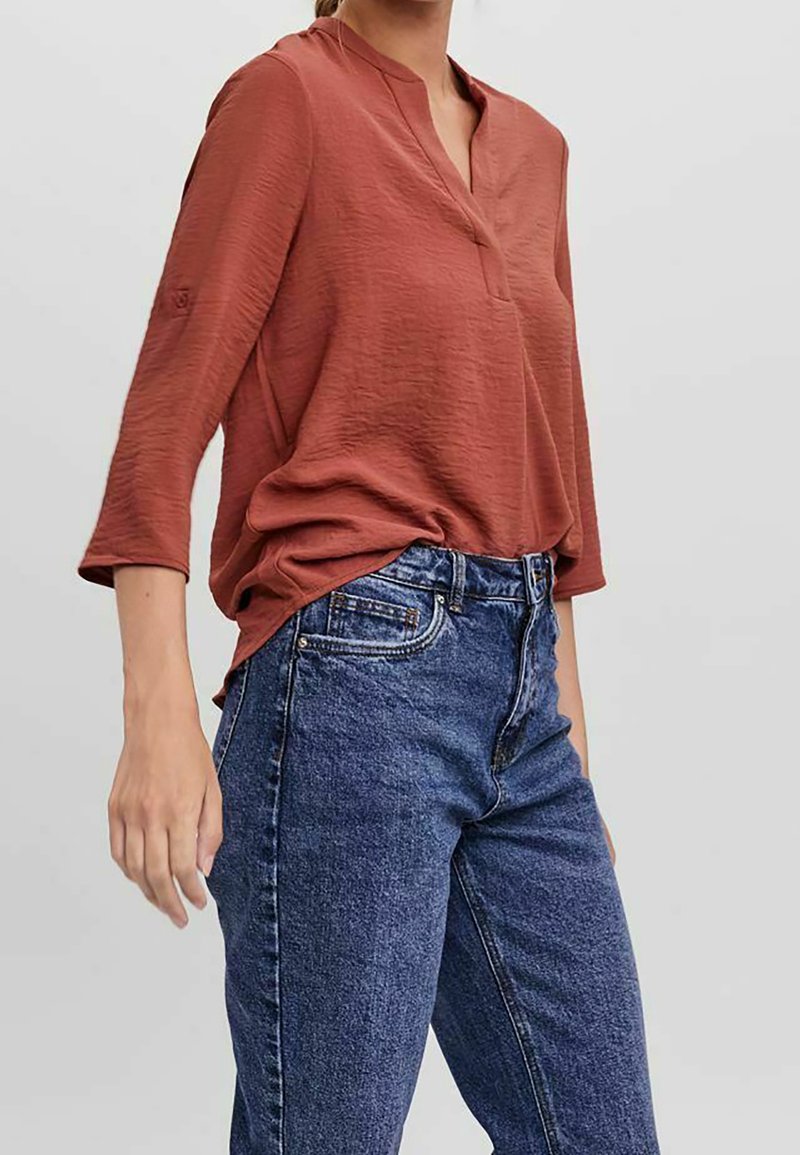 Femme portant un chemisier à manches longues de couleur rouille, rentré dans un jean en denim bleu taille haute, sur un fond uni.