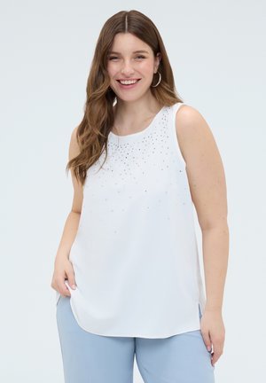 Smilende kvinde med langt brunt hår iført sølv øreringe i hoops, en hvid ærmeløs top med rhinsten og lyseblå bukser mod en ensfarvet baggrund.