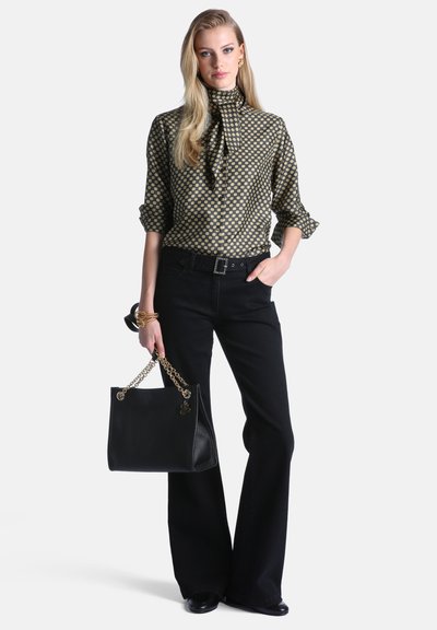 Femme portant un chemisier à motifs et un pantalon noir à jambes larges tenant un sac à main noir avec des anses en chaîne dorée.