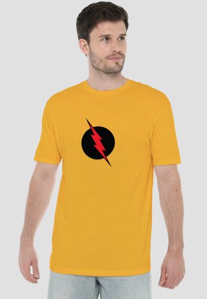 Mann trägt ein gelbes T-Shirt mit schwarzem Kreis und rotem Blitzdesign, schaut nach links.