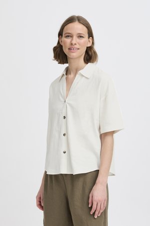 Jeune femme aux cheveux bruns moyens portant une chemise blanche à manches courtes, ample et boutonnée, ainsi qu'un pantalon vert olive, sur un fond uni.
