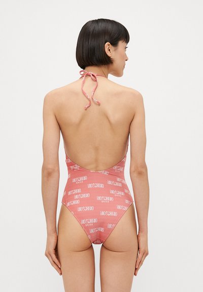Maillot de bain rose avec une taille haute, un dos nu, un nœud au cou et un motif répétitif du logo "MOSCHINO" en blanc sur l'ensemble du tissu.