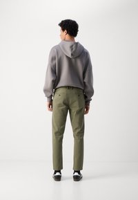 Hanorac gri cu glugă mare, croială supradimensionată și manșete strâmte; asortat cu pantaloni chino de culoare verde măsliniu și adidași negri cu alb.