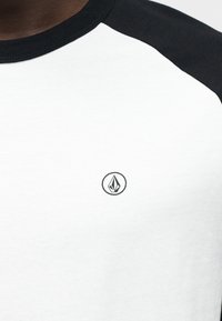 Μαύρο και λευκό T-shirt με στρογγυλή λαιμόκοψη, με λευκό σώμα και μαύρα ραγκλάν μανίκια. Ένα μικρό μαύρο λογότυπο βρίσκεται στο στήθος.