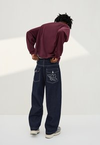 Persoon in donkergroene wijde spijkerbroek met sterpatch op zak en bordeauxrode longsleeve die de spijkerbroek optrekt of aanpast.