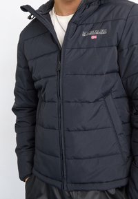 Kurtka puchowa NapaPuri w kolorze granatowym, z pikowanym wzorem, zamkiem błyskawicznym oraz naszywką z logo i małym detalem w postaci flagi. Tkanina ma gładką teksturę.