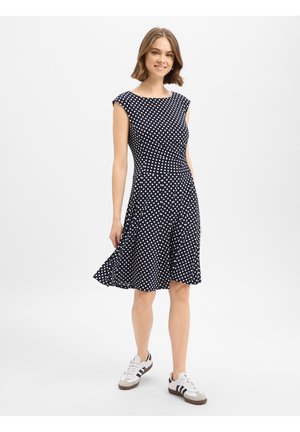 Junge Frau lächelt, trägt ein ärmelloses dunkelblaues Kleid mit weißen Punkten und weiße Turnschuhe mit schwarzen Streifen, steht auf weißem Hintergrund.