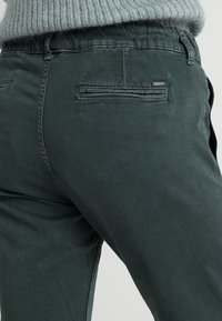 Pantalones casuales de color verde oscuro con bolsillos traseros de ojal y cinturilla elástica, combinados con un suéter de punto en gris claro.