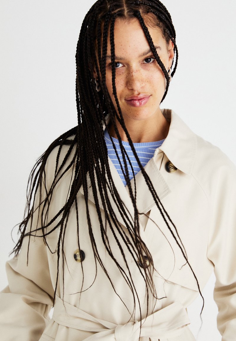 Beżowy płaszcz trench z paskiem w talii, guzikami podwójnego rzutu i klasycznym kołnierzem. Noszony na jasnoniebieską bluzkę w paski.