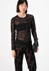 Zwarte mesh lange mouw top met ingewikkelde zwarte geborduurde patronen, gecombineerd met bijpassende zwarte gestructureerde broek en een elegante zwarte clutch.