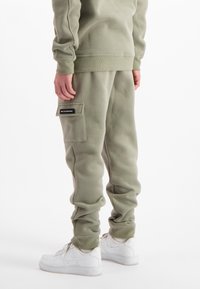 Persoon in olijfgroene cargo-sweatpants met een zijzak en witte sneakers, staand tegen een witte achtergrond.