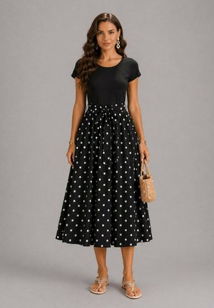 Femme portant un haut noir à manches courtes et une jupe midi noire taille haute à pois blancs, tenant un sac à main tissé, debout sur un fond gris.