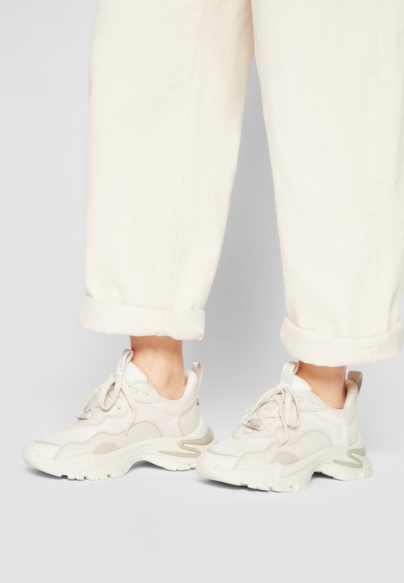 Světlé béžové sportovní tenisky z prodyšného a syntetického materiálu; kulatá špička, silná podrážka a tkaničky; spárované s ohrnutými krémovými kalhotami.