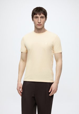 Čovjek u blijedo žutoj majici kratkih rukava i tamnosmeđim hlačama stoji nasuprot jednobojnoj svijetloj pozadini.