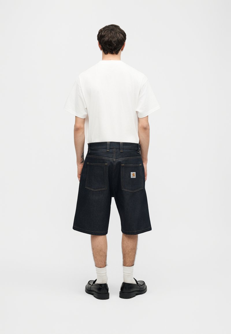 Uomo con maglietta bianca, pantaloncini di jeans scuri fino al ginocchio con tasche posteriori, calzini bianchi e scarpe nere, di spalle su sfondo semplice.