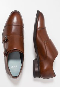 Zapatos de cuero marrón con un acabado suave, doble hebilla, puntera redondeada y un tacón bajo apilado; el interior presenta una plantilla de color azul claro.