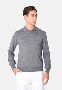 Polo grigio a maniche lunghe con tre bottoni, tessuto testurizzato, colletto standard e polsini a coste. Indossato con pantaloni bianchi.