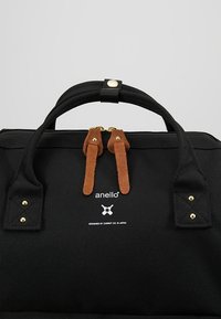 Sac Anello noir avec double poignée, tirettes de fermeture éclair en daim marron, quincaillerie dorée et texte du logo blanc au centre avant.
