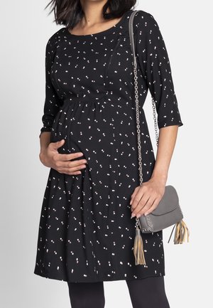 Femme enceinte portant une robe noire à petits pois blancs et roses, tenant son ventre d'une main, portant un sac gris à franges.